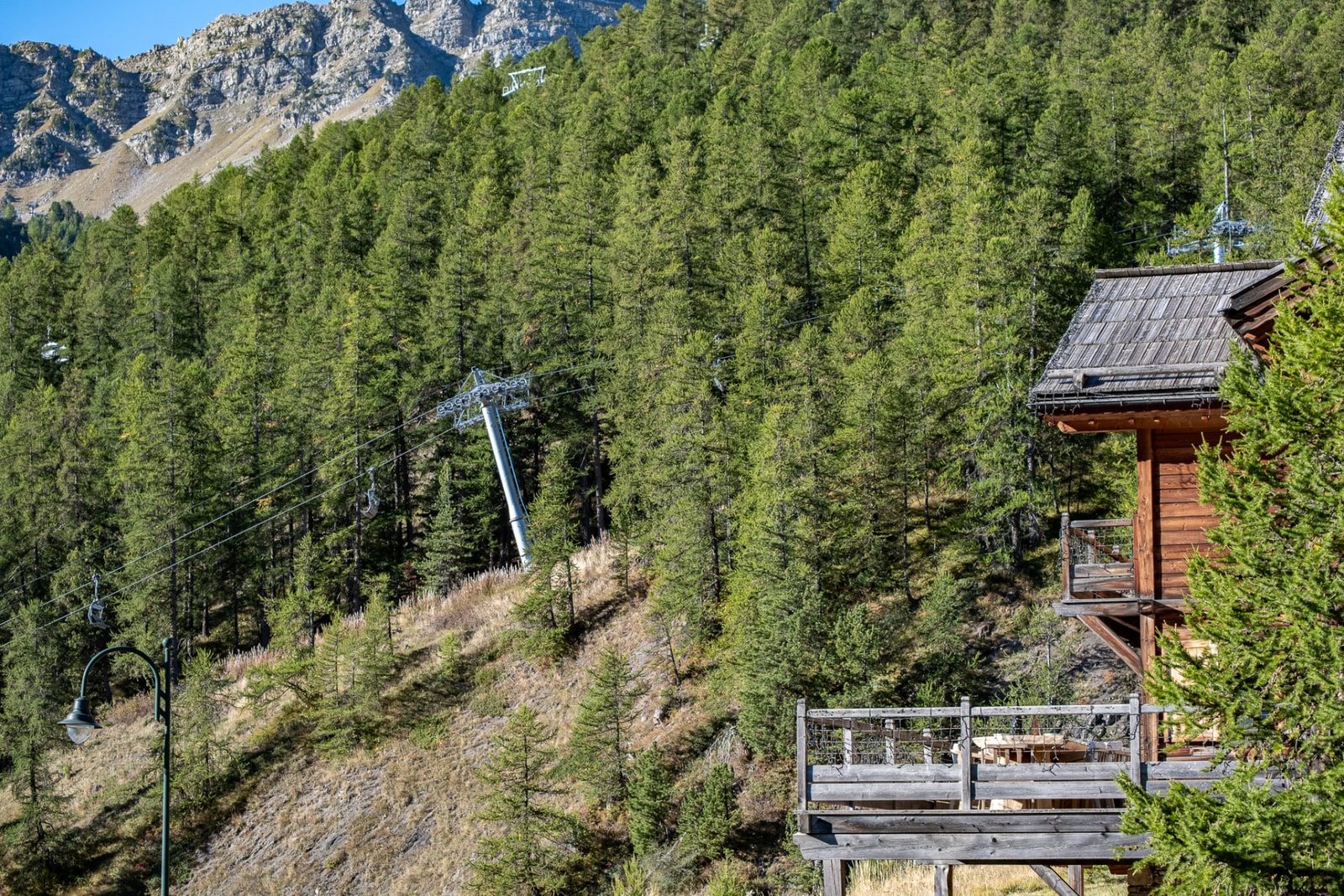 Chalet en bois et sa terrasse au pied d'une pente boisée traversée par un télésiège, sous les sommets de Vars