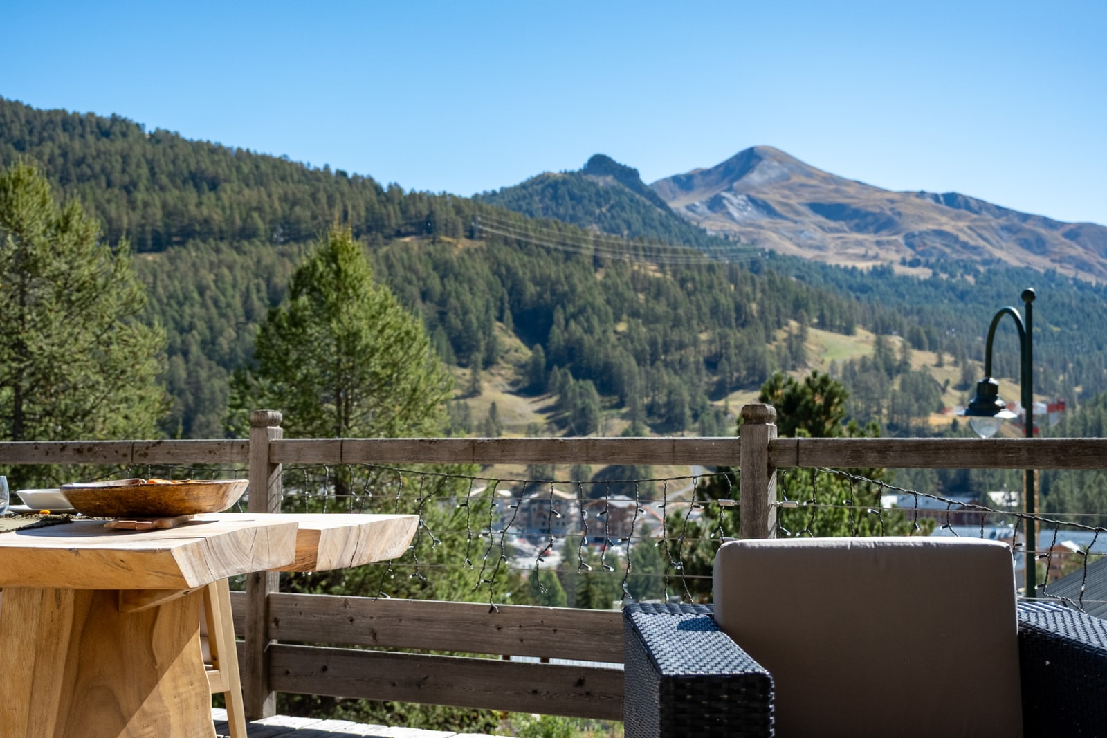 Terrasse en bois avec une table brute et un fauteuil, face aux pentes boisées et à un sommet sous un ciel bleu